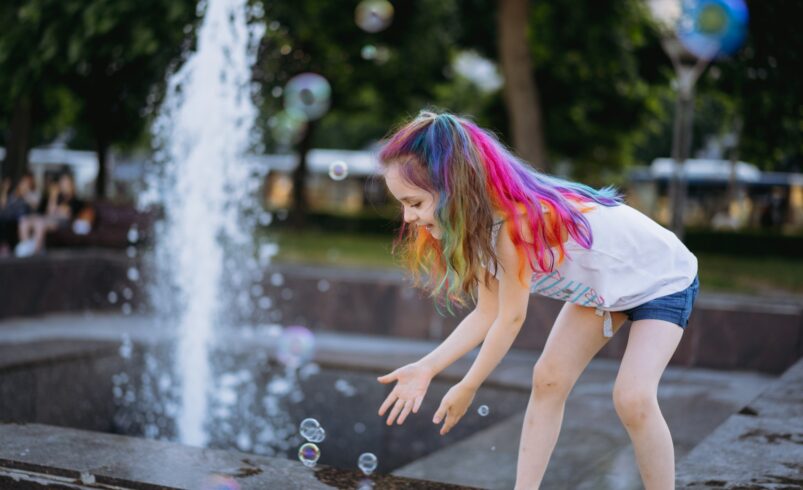 cute little girl with colorful hair catchilng soap bubbles by splashing fountain in city park